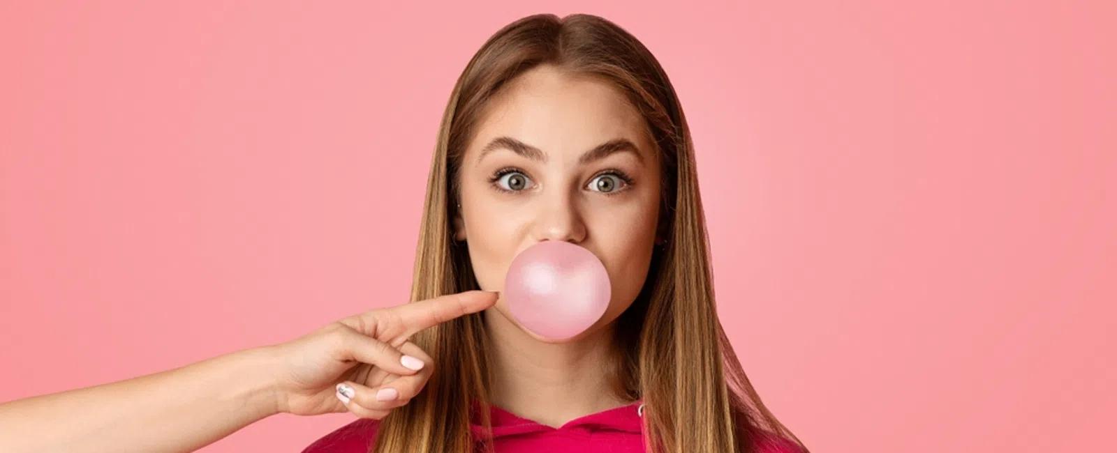 Young woman blowing a pink bubblegum bubble with a finger about to pop it against a pink background.