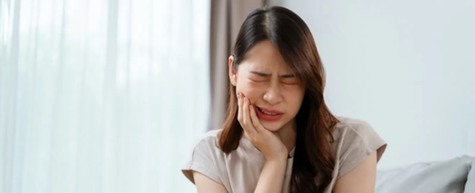 Young woman sitting on couch holding her cheek in pain showing toothache or jaw discomfort.