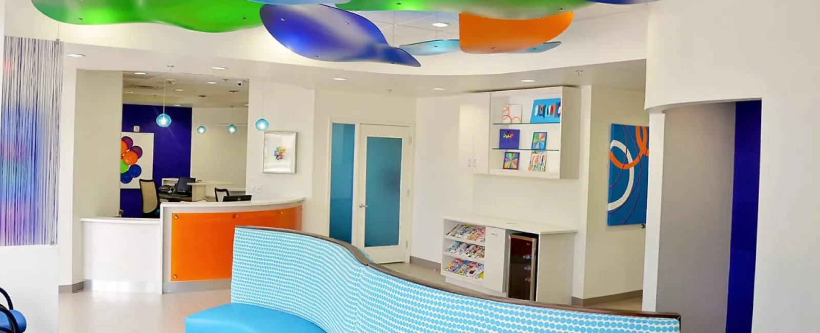 Modern dental office waiting area with colorful ceiling panels, blue curved bench, and bright reception desk.
