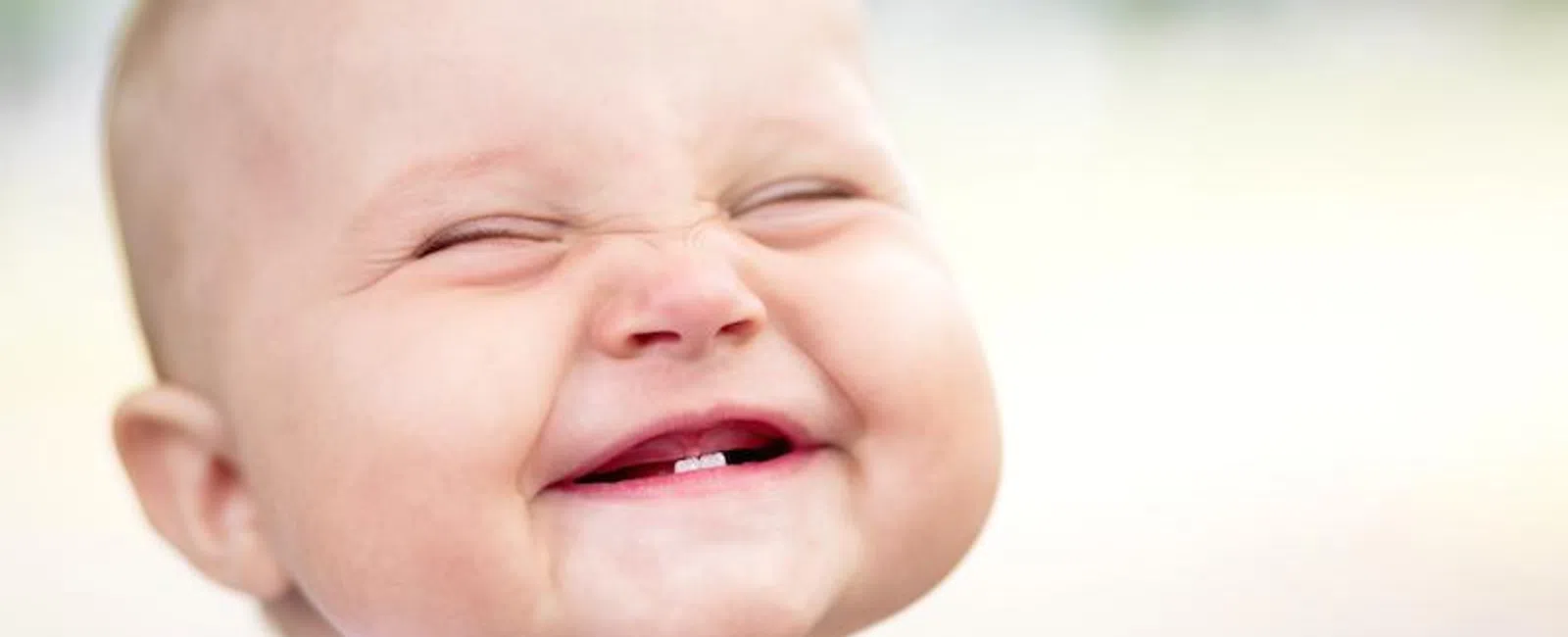 Close-up of a happy baby smiling with eyes closed showing two baby teeth, blurred background.