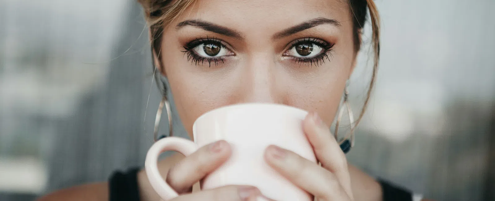 Close-up of a woman with expressive eyes holding and drinking from a white mug indoors.