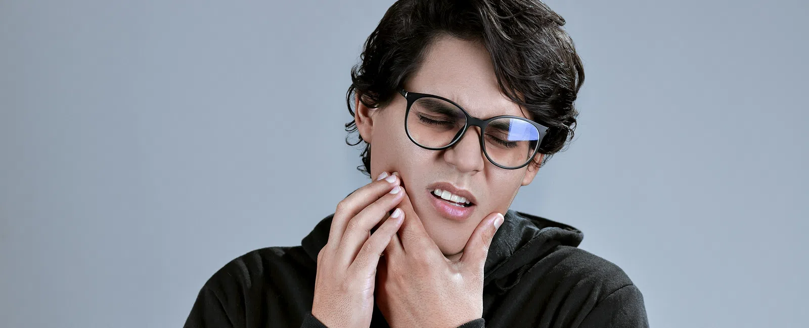 Young man in glasses wearing black hoodie, holding jaw in pain, showing toothache or dental discomfort