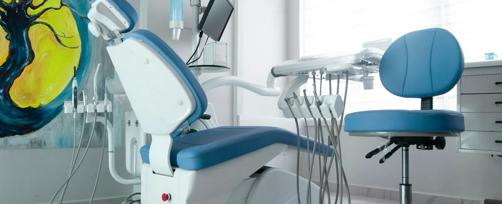 Modern dental clinic interior with blue dental chair, equipment, and colorful tree artwork on the wall.