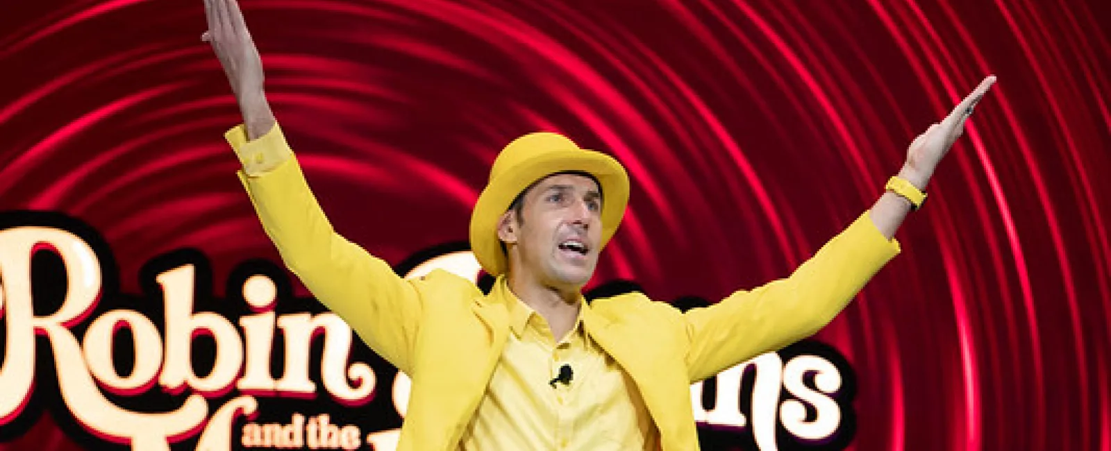 Man in bright yellow suit and hat gestures with arms raised on stage with Robin Williams tribute backdrop.