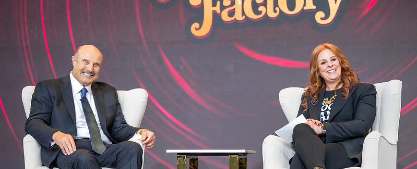 Man and woman seated on white chairs on stage during a conversation event with a table between them.