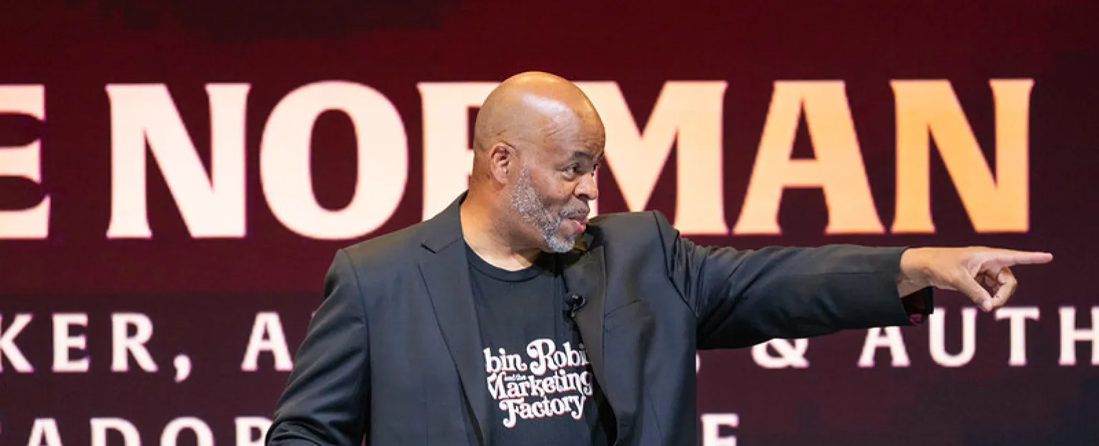 Bald man in black blazer and graphic t-shirt gestures and points during a presentation on stage.