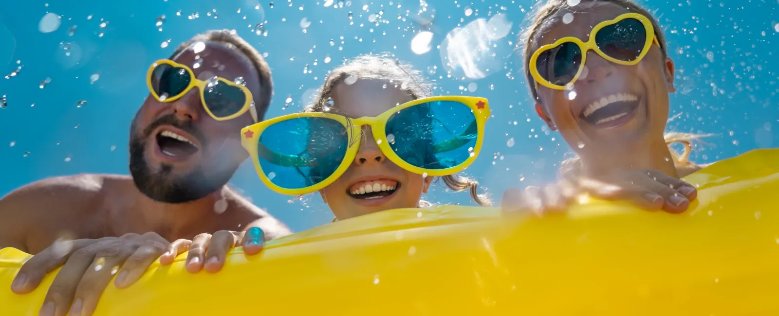 Three smiling people wearing large colorful sunglasses enjoying a sunny day on a yellow pool float splashing water.