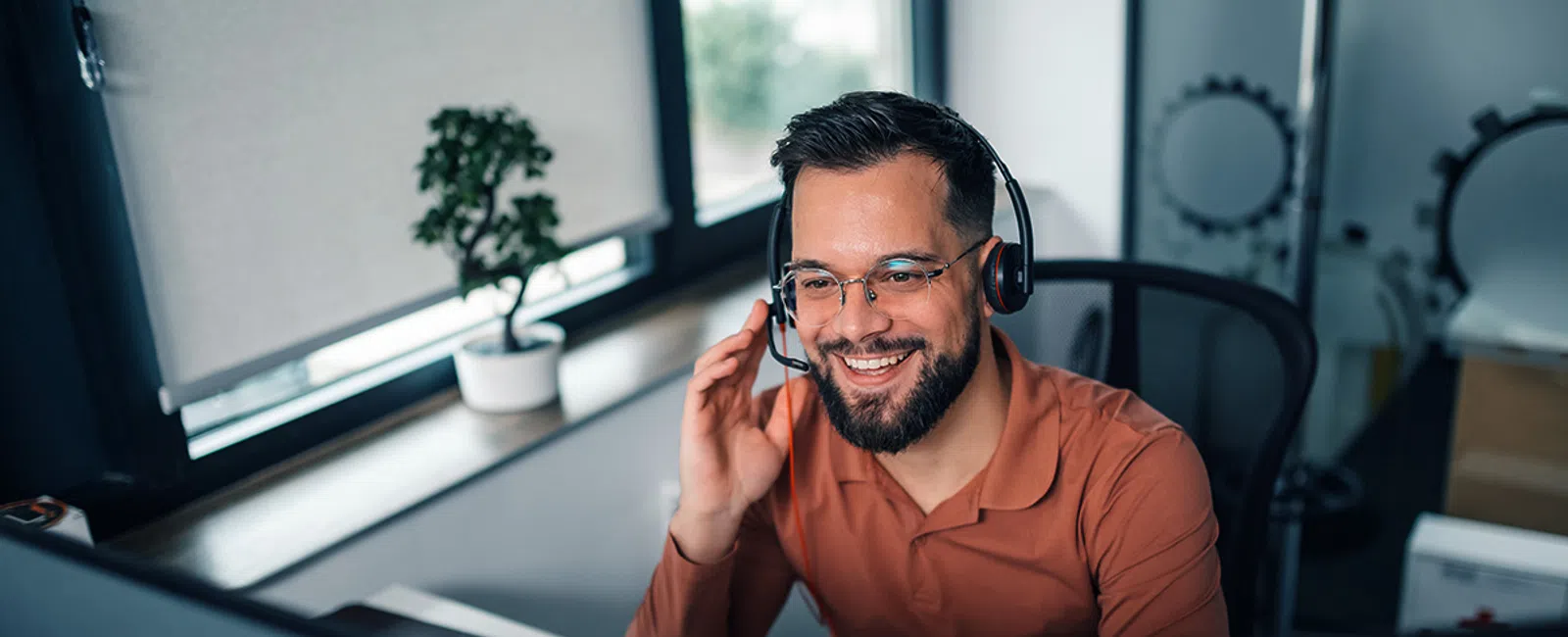 Smiling man wearing headset working at dual monitors in modern office with natural light and indoor plants