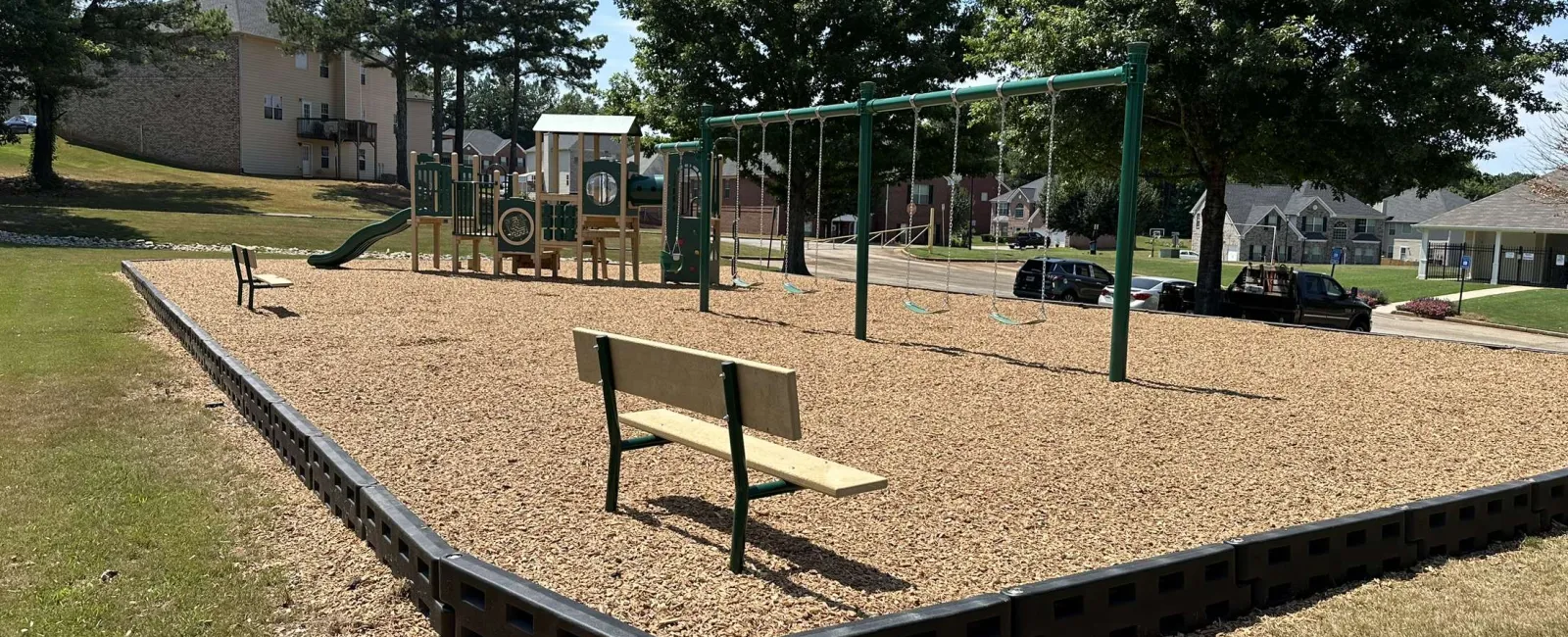 Outdoor playground with swings, slide, benches, and wood chip ground covering on a sunny day.