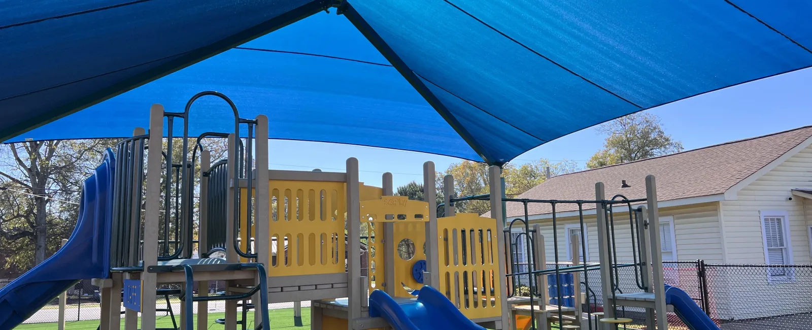 Children's playground with blue slides and yellow climbing structures under a large blue shade canopy.
