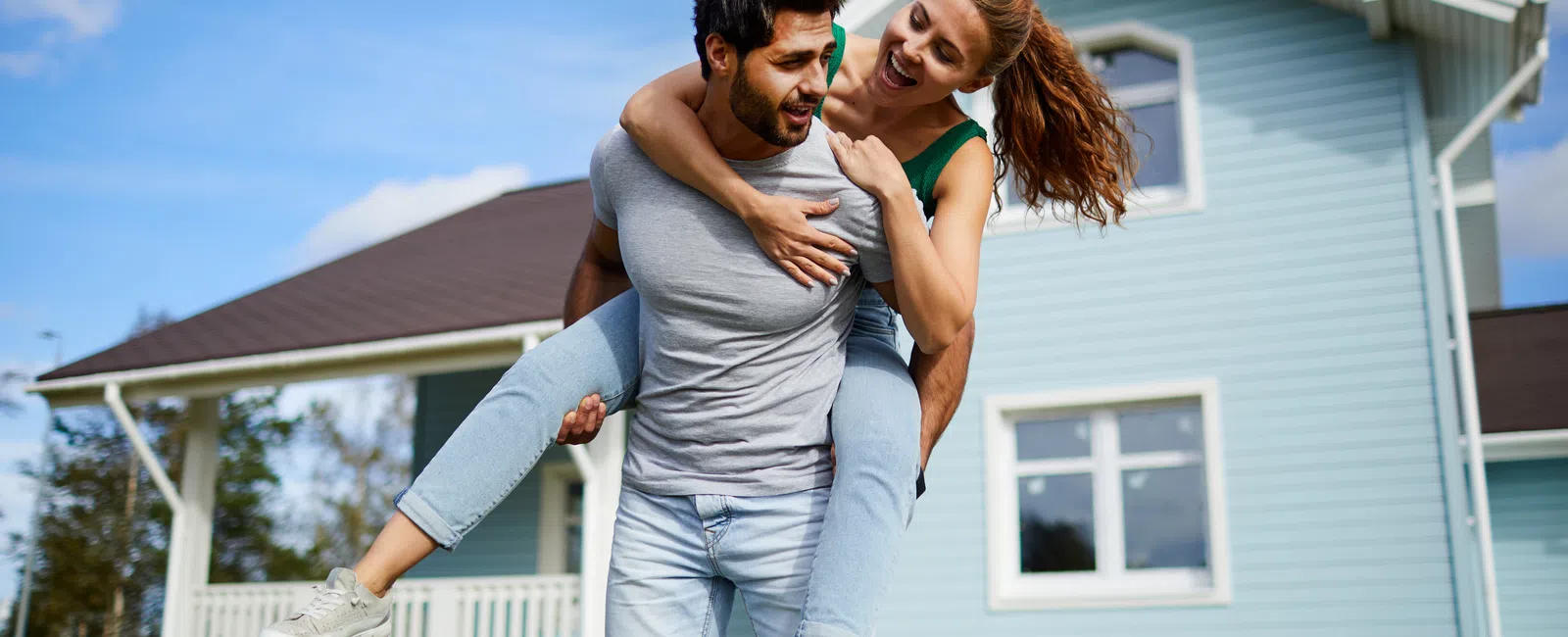 Happy young couple with woman piggyback on man in front of blue suburban house on sunny day