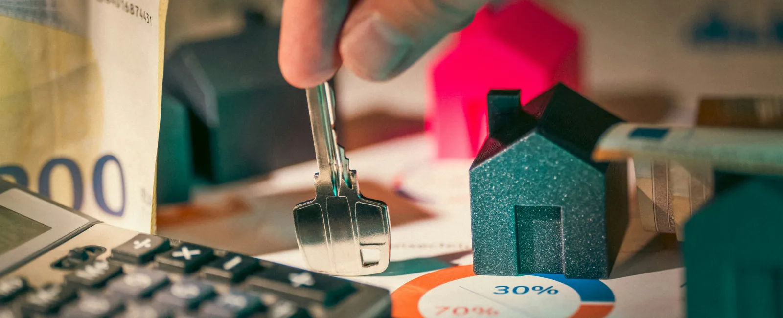Hand holding a key over house models, calculator, cash, and pie chart representing real estate investment and mortgage finance.