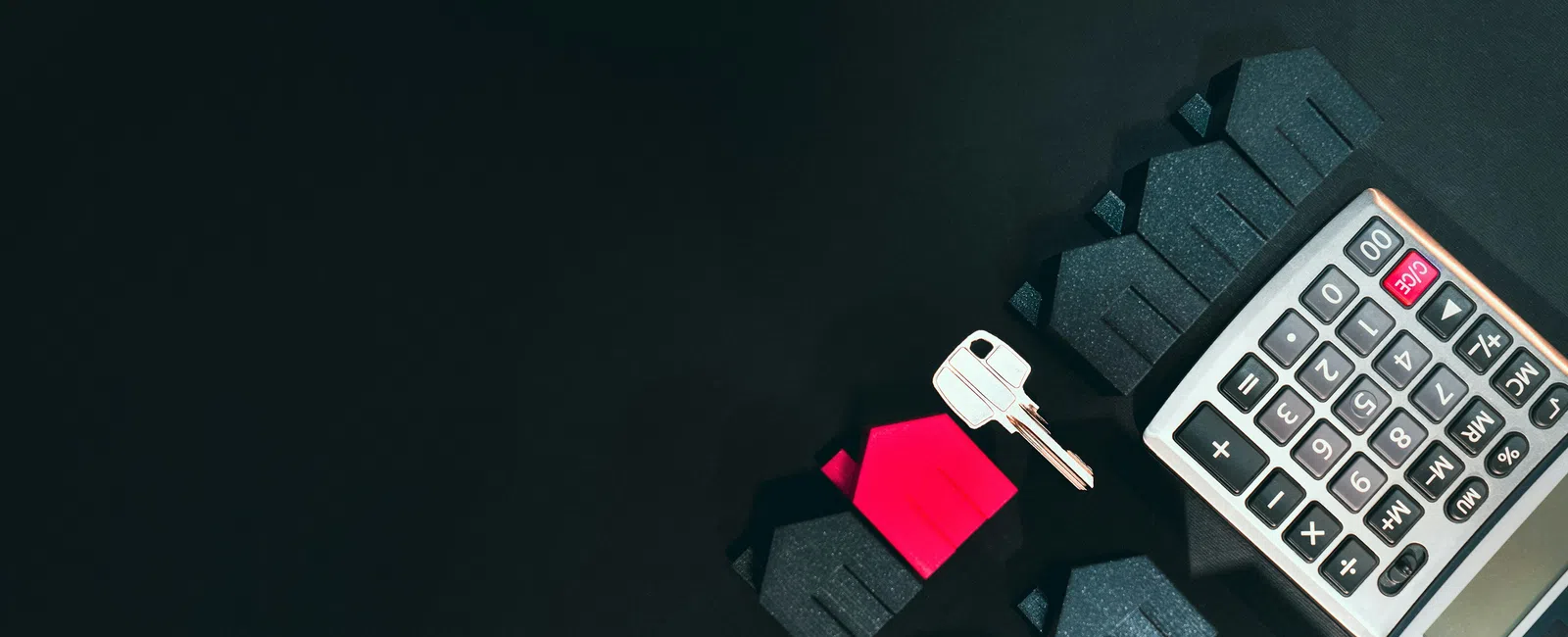 Calculator, house-shaped figures in black and red, and a silver key on a dark surface representing real estate finance.