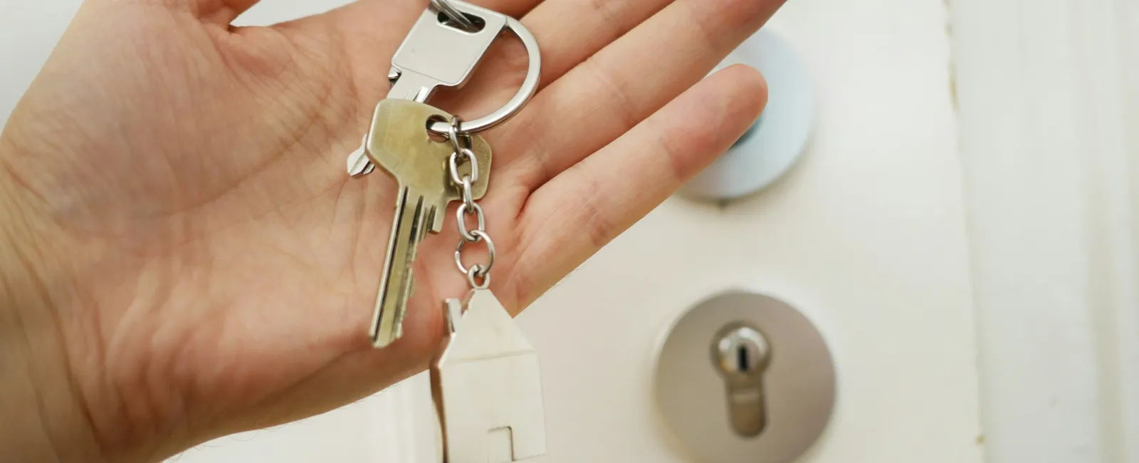 Hand holding house-shaped keychain and keys near a door lock symbolizing home ownership or real estate.