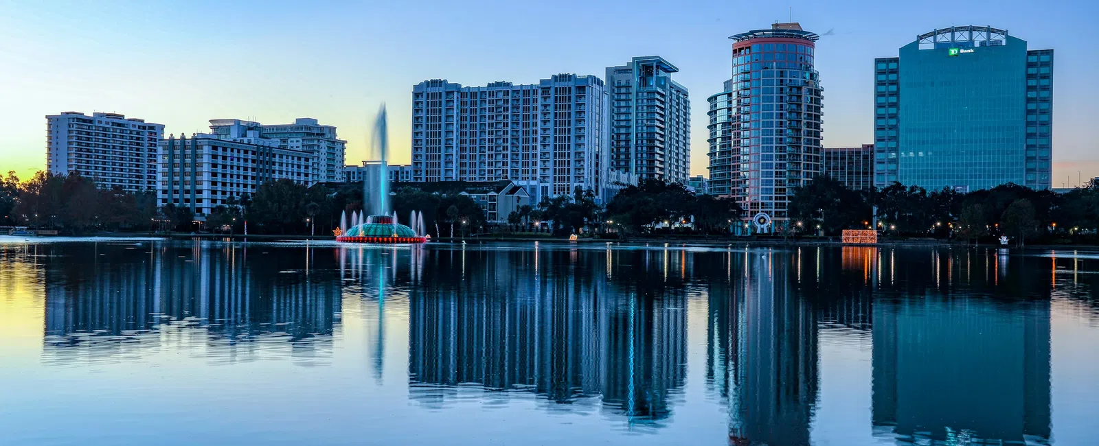City skyline with tall buildings and central fountain reflecting on calm lake water at dusk.
