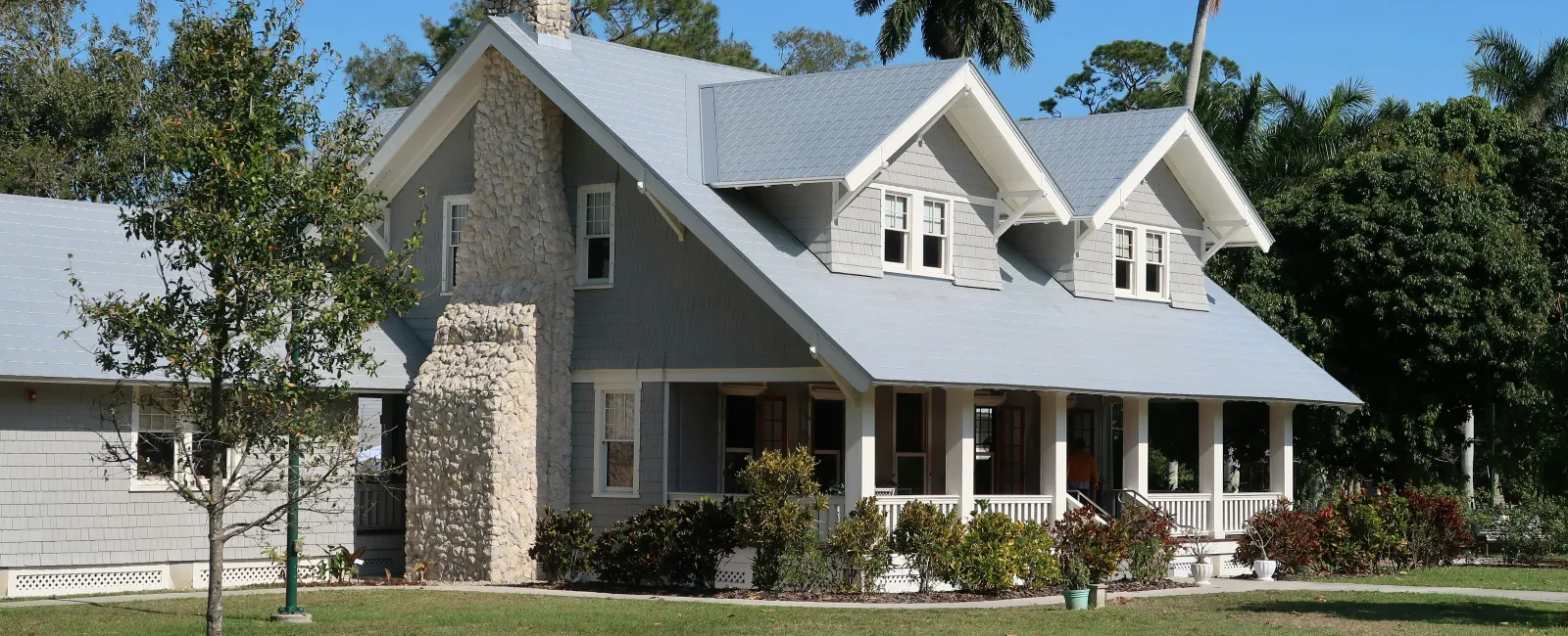 Gray craftsman-style house with stone chimney, large porch, and lush green lawn under clear sky.