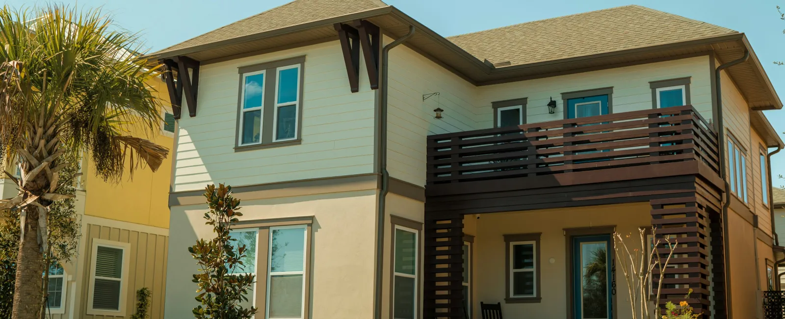 Two-story beige and cream house with wooden balcony and front porch in a sunny residential neighborhood