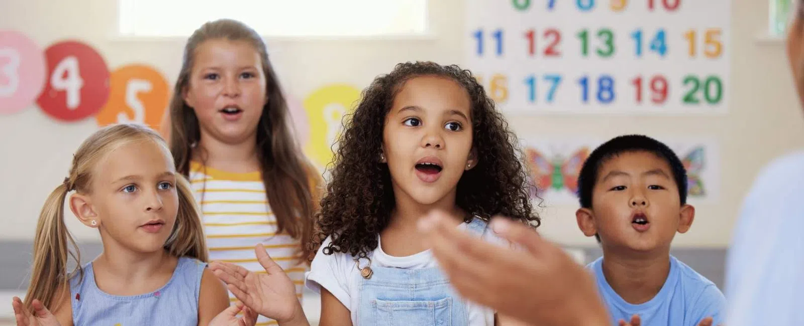 Diverse group of children clapping and singing in a colorful classroom setting with a teacher.
