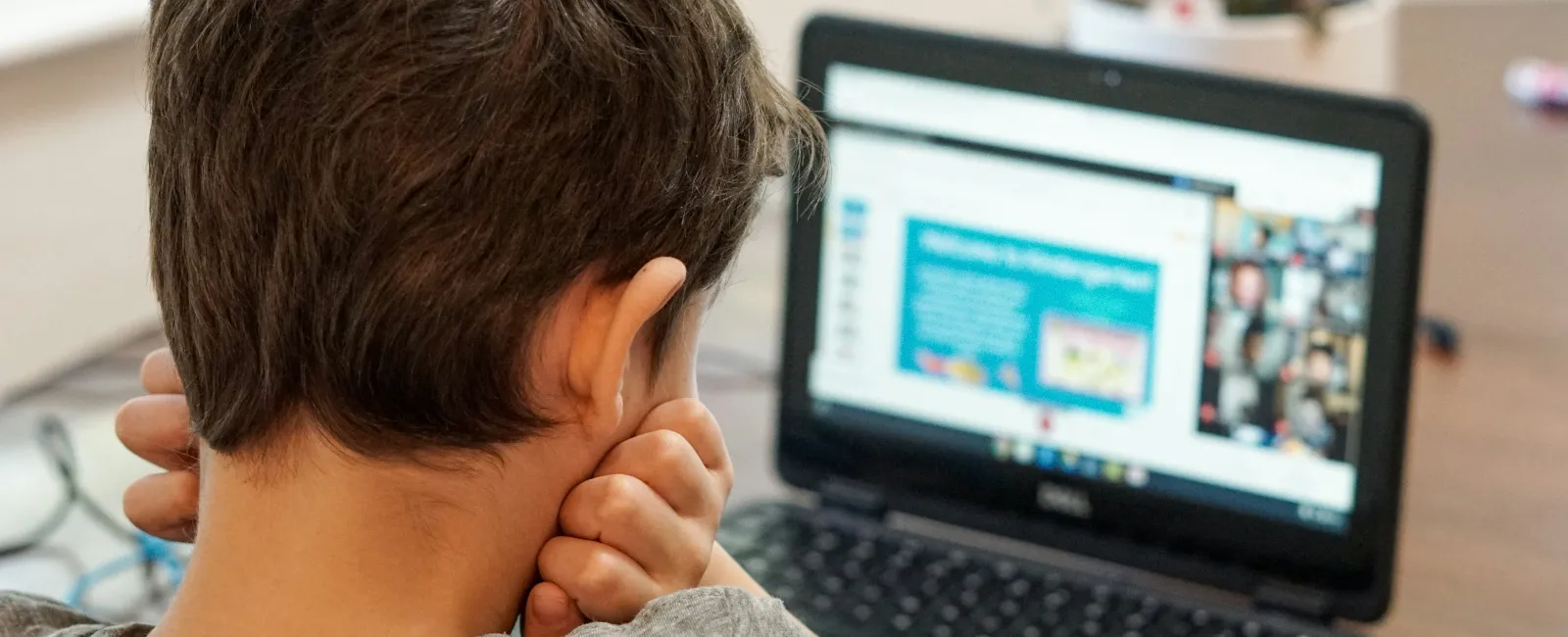 Child sitting at a table watching an online class on a laptop with a blurred screen and video call participants.