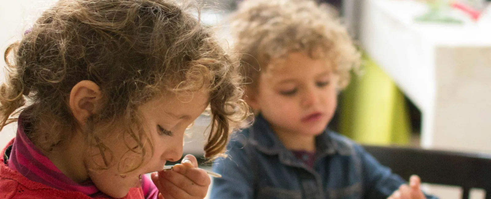Two young children eating fruit at a wooden table while an adult prepares food in the kitchen background