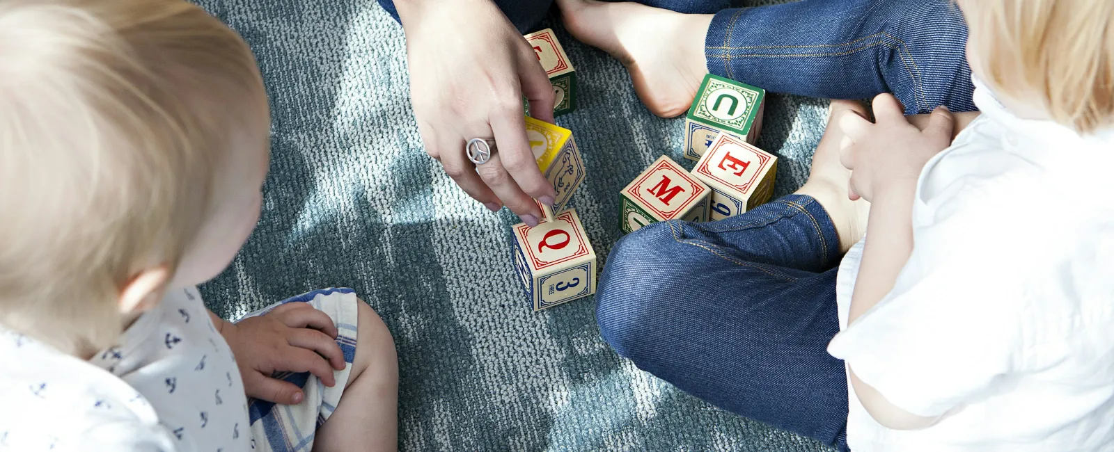 Two toddlers and an adult sitting on carpet playing with wooden alphabet blocks together.