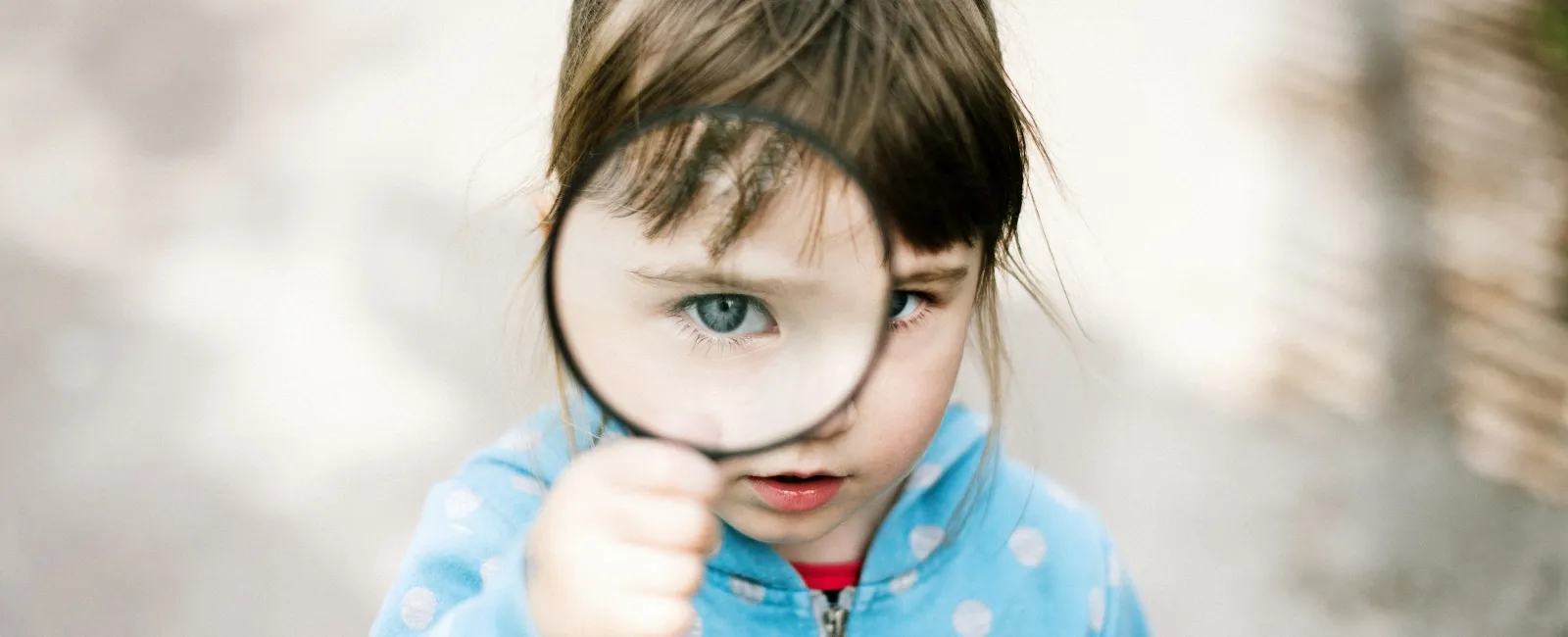 Young girl in blue polka dot jacket holding magnifying glass to her eye outdoors