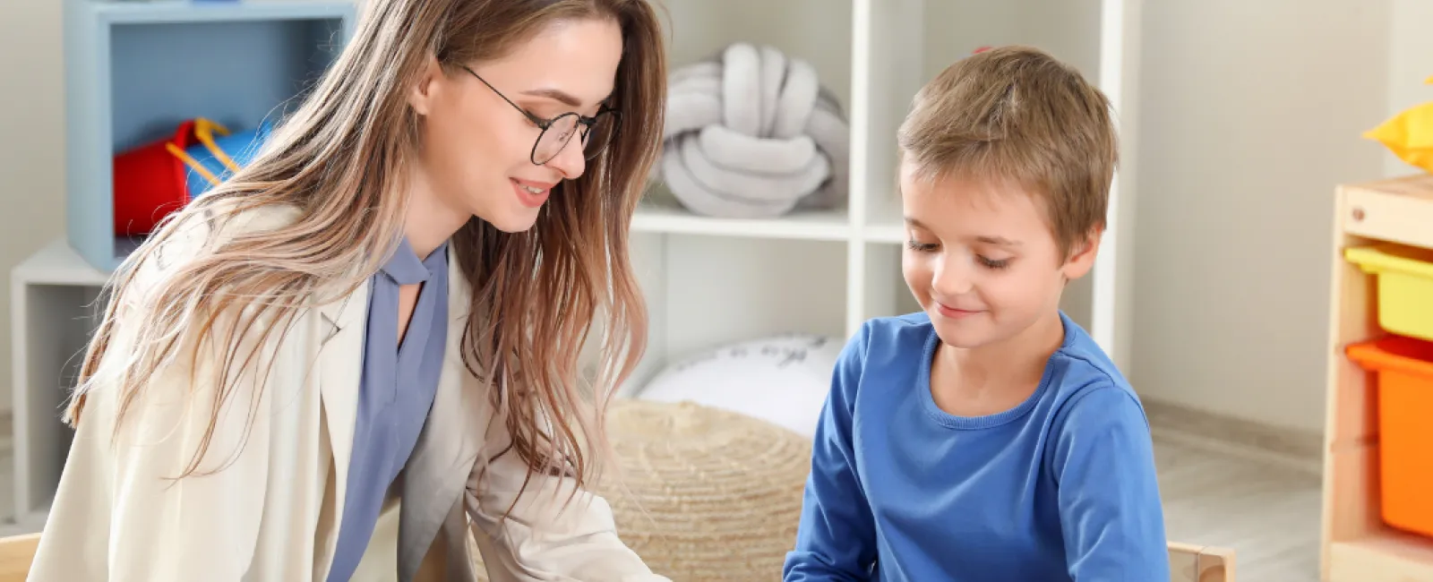 Young woman helps boy learn with colorful alphabet letters on a table in a bright playroom.
