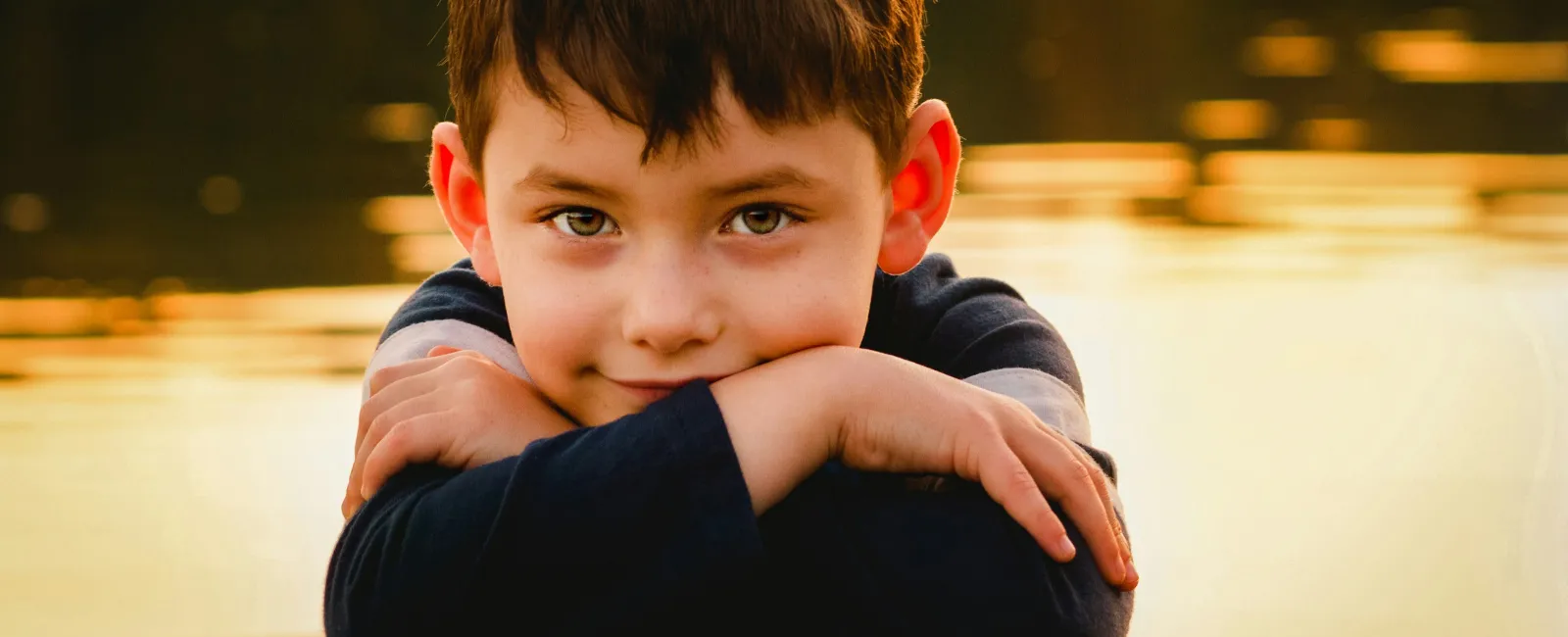 Young boy with brown hair resting arms on a surface by golden water at sunset, smiling gently at camera.