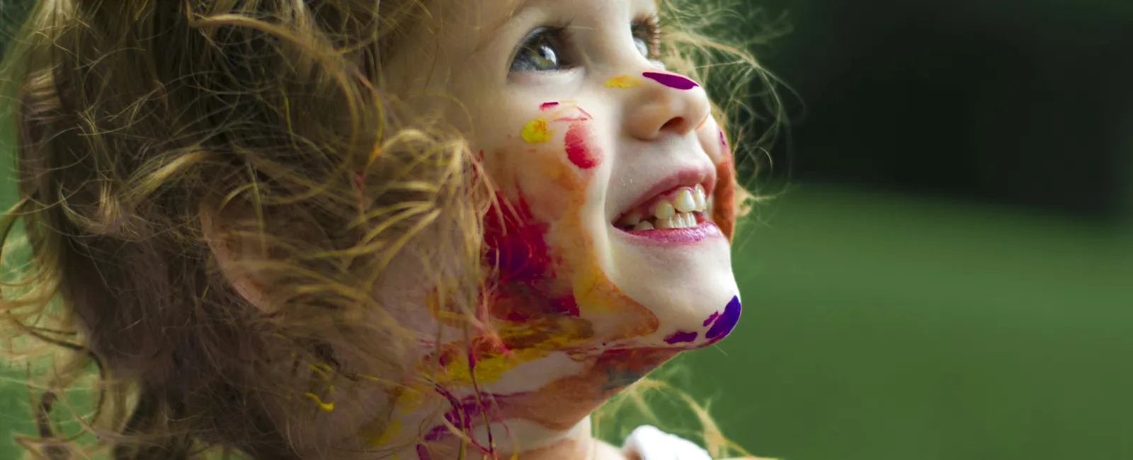 Smiling toddler girl with curly hair and yellow bow covered in colorful paint outdoors on a green background.
