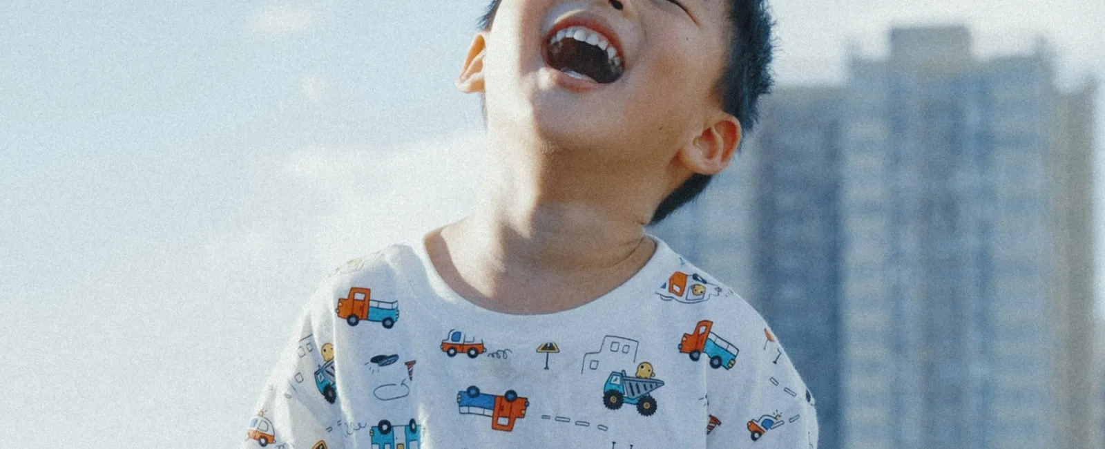 Smiling young boy wearing a colorful vehicle print t-shirt and shorts outdoors on a sunny day with buildings in background