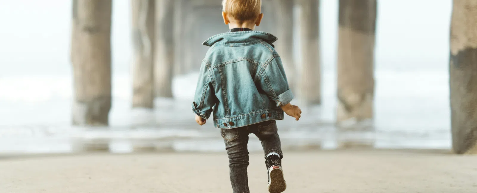 Child in denim jacket walking on sandy beach under pier pillars near the ocean shore
