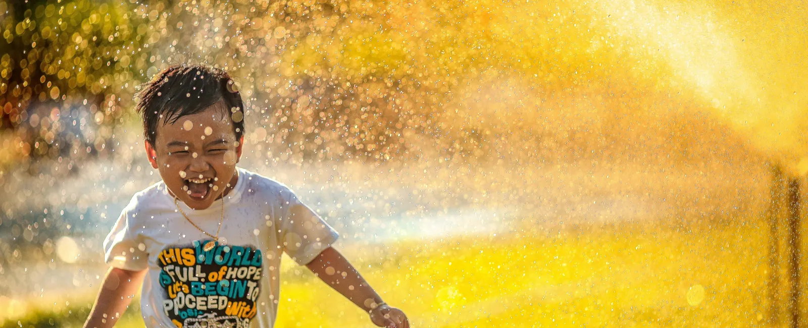 Happy child running through a water sprinkler on a sunny day with bright golden light and water droplets.