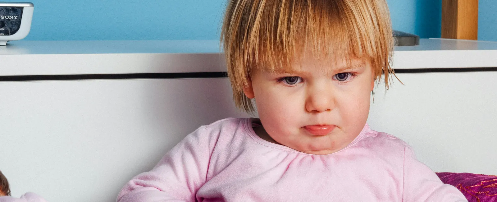 Young child with blond hair wearing pink pajamas sitting on bed with a serious expression.