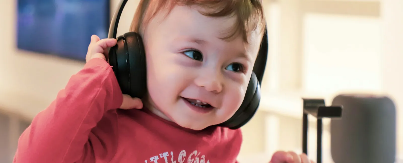 Smiling toddler wearing black headphones and a red shirt playing indoors in a bright room.