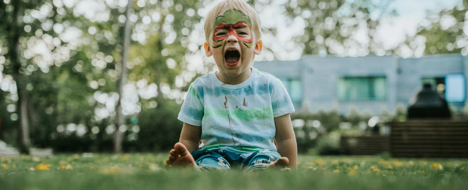 Young child with butterfly face paint sitting on grass with mouth wide open outdoors on a sunny day