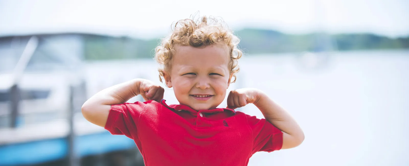 Smiling curly-haired boy in a red shirt flexes muscles near a lakeside dock on a sunny day