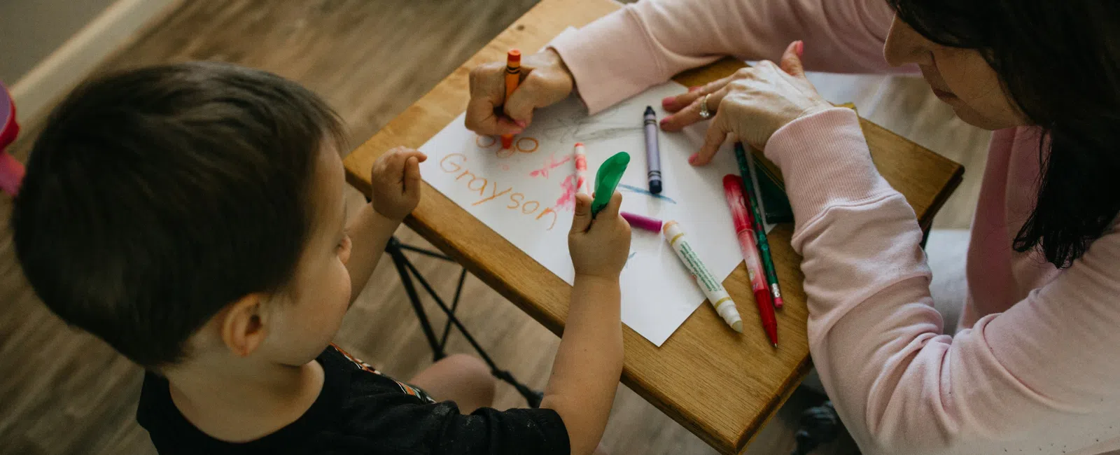 Young child and adult drawing with crayons and markers at a small wooden table indoors.