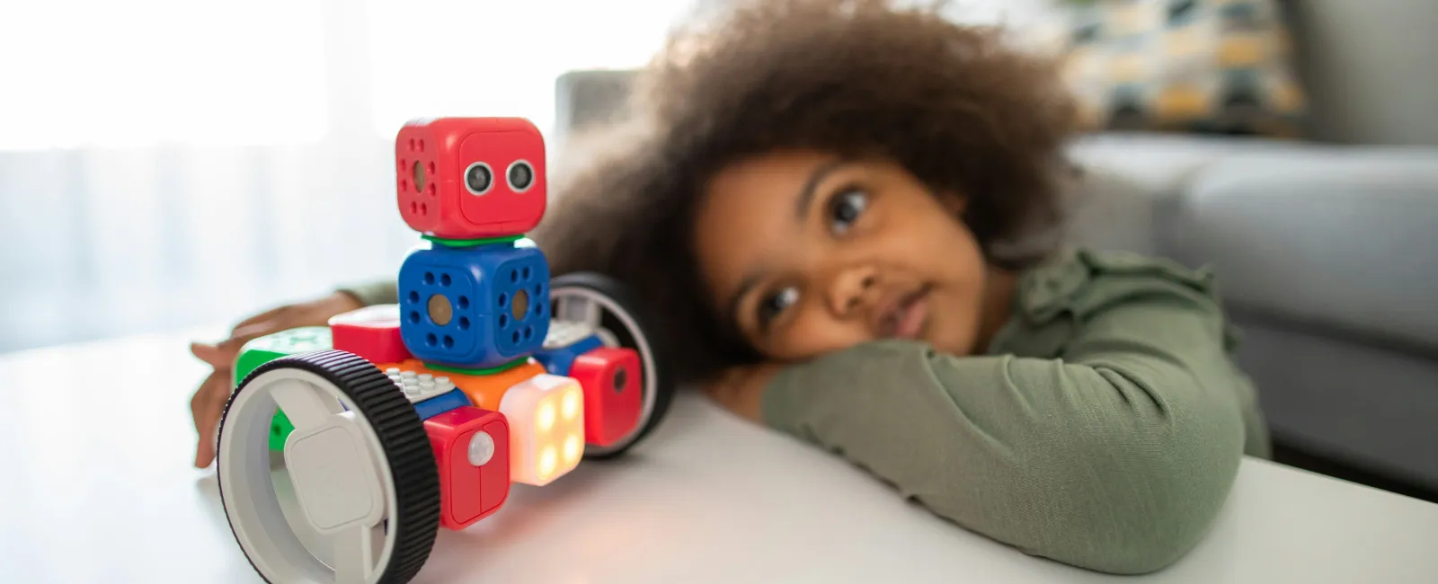 Curious child watches colorful robotic toy with large wheels and glowing lights on white table indoors.
