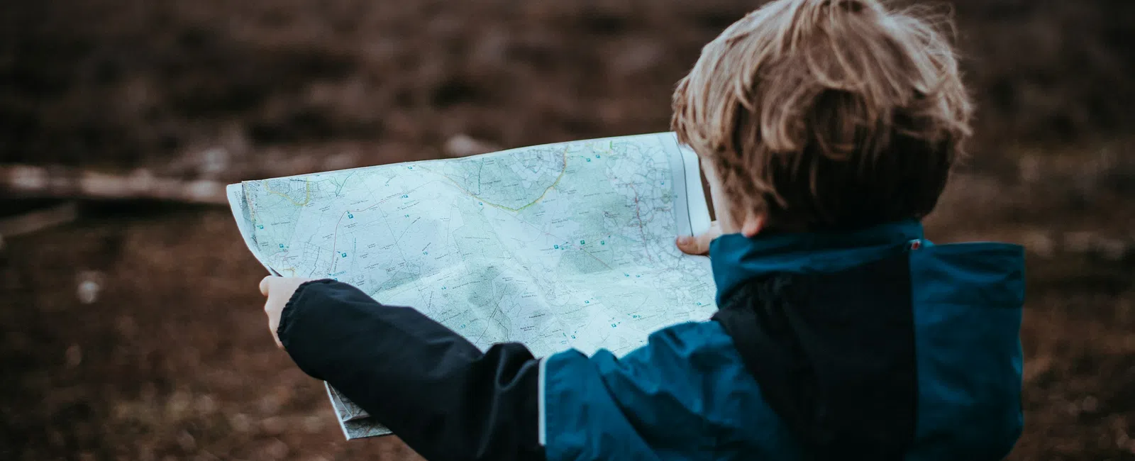 Child wearing blue jacket studies map outdoors in a brown barren landscape under cloudy sky.