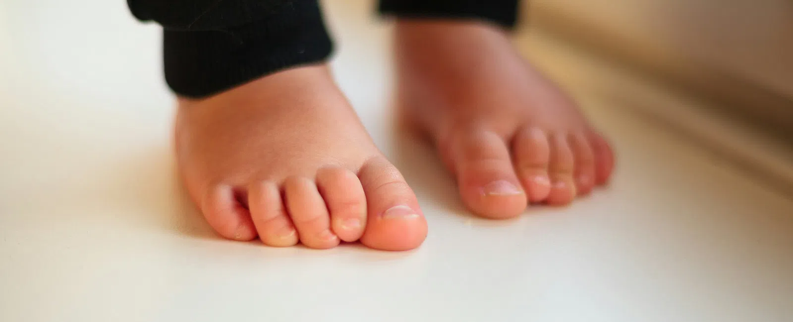 Close-up of a toddler's bare feet standing on a white surface wearing black pants.