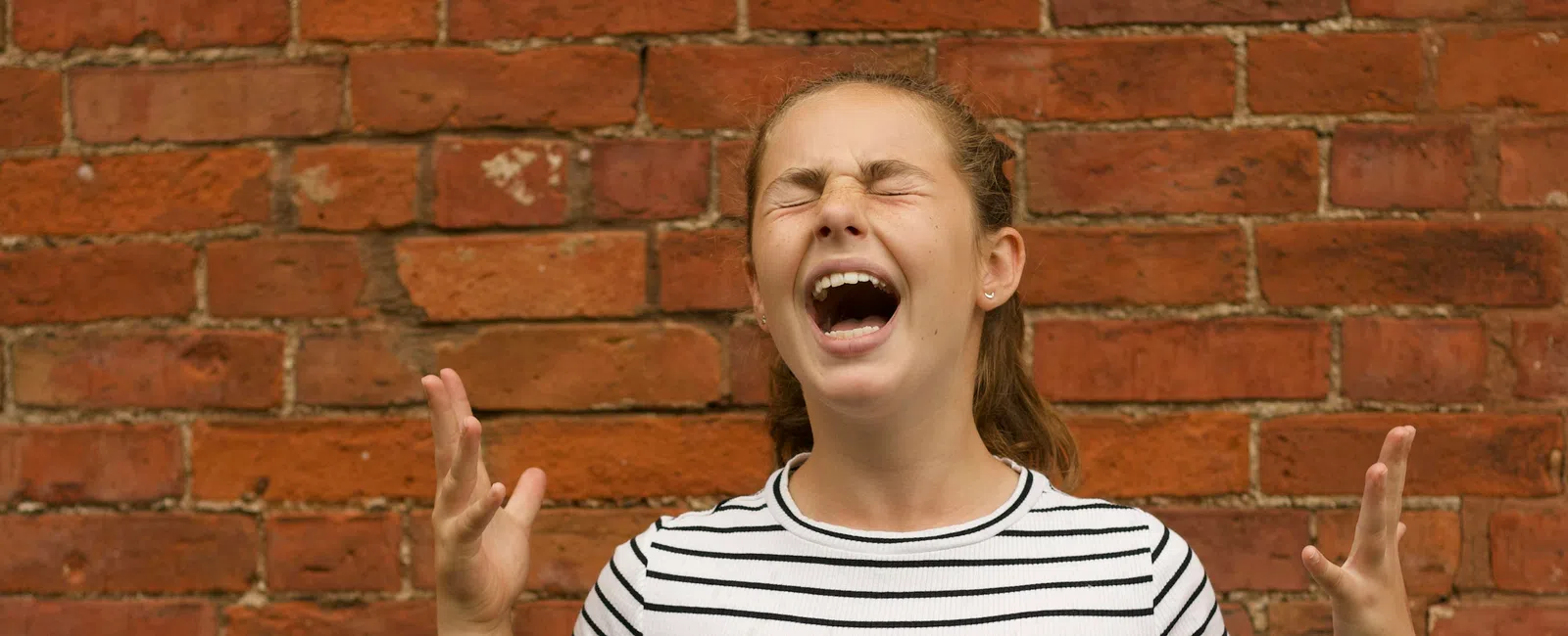 Young woman with eyes closed and hands raised, expressing frustration in front of a red brick wall.