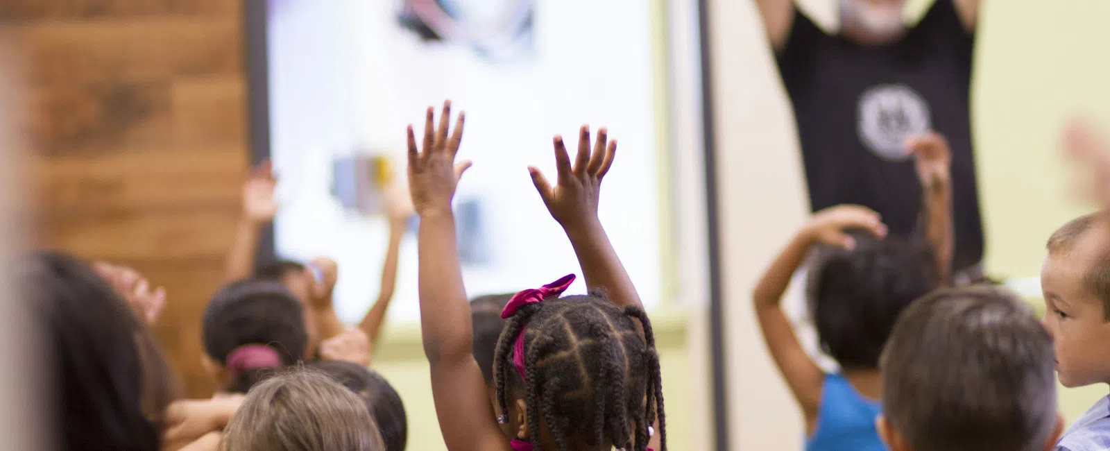 Children raising their hands participating in an activity led by an instructor in a classroom setting.