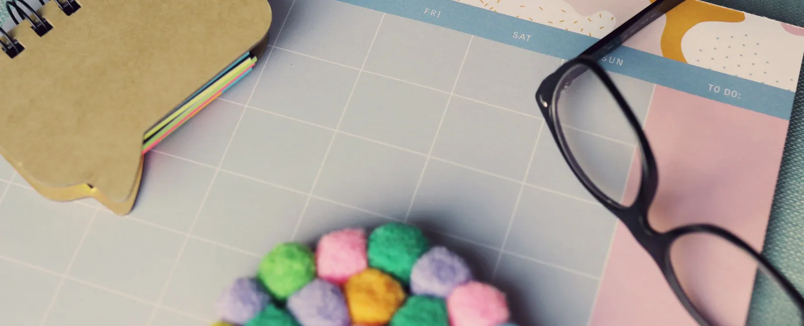 Weekly planner with colorful sticky notes, round pom-pom coaster, and black eyeglasses on blue surface.