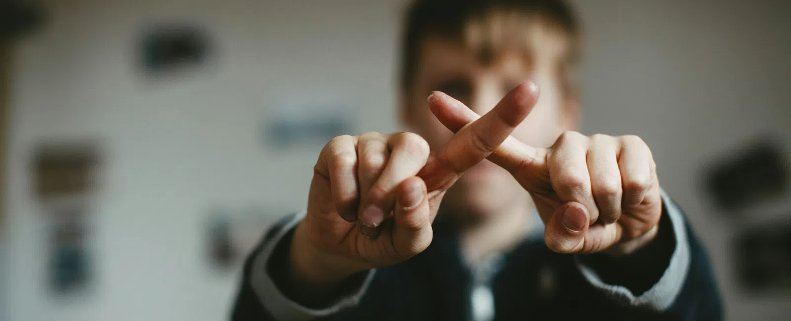 Close-up of a child crossing fingers to form an X sign with blurred background indoors.