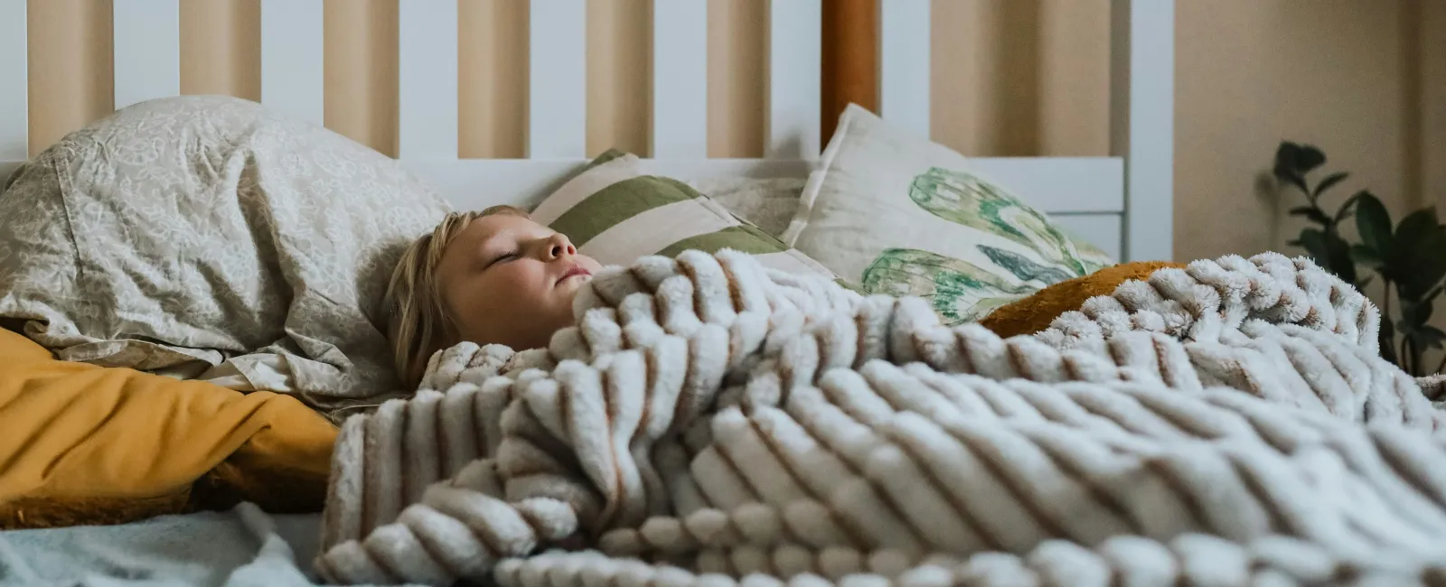 Young child sleeping peacefully under a soft, chunky knit blanket on a cozy bed with pillows.