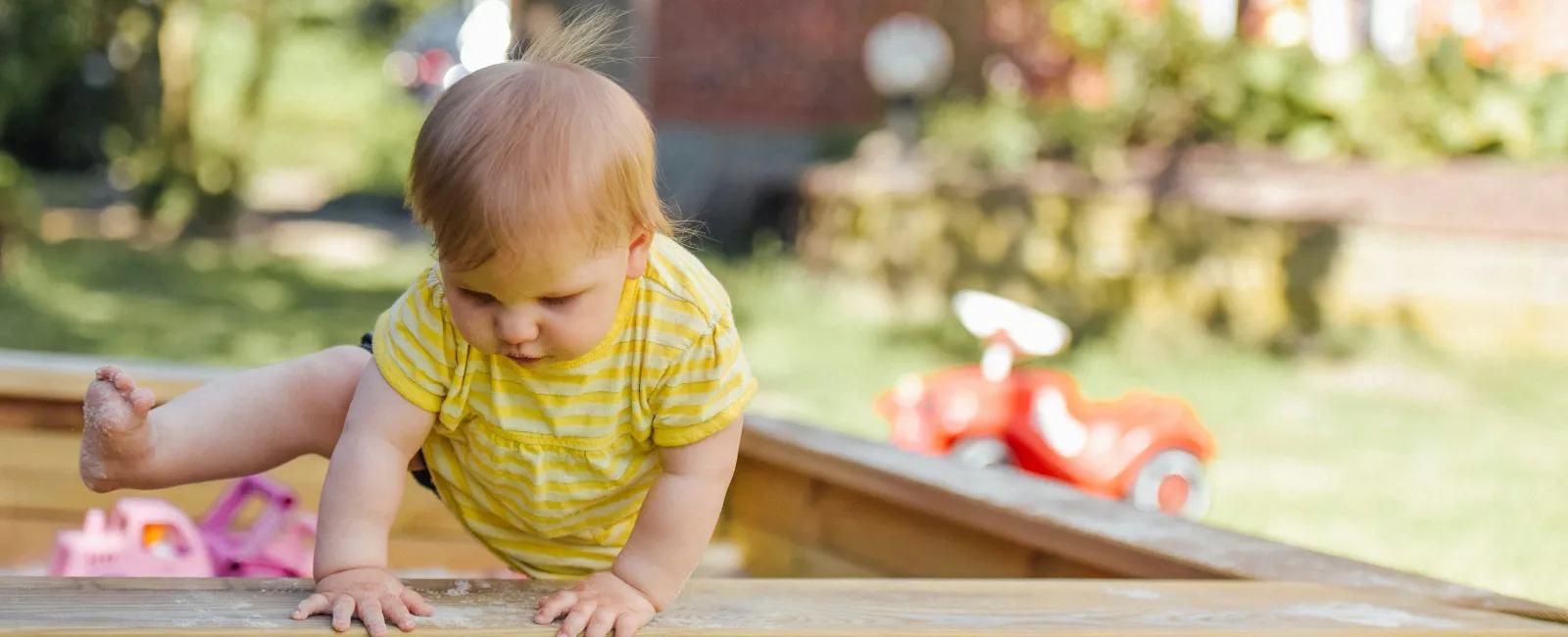 Toddler in yellow outfit climbing out of a wooden sandbox in a sunlit backyard with toys around.