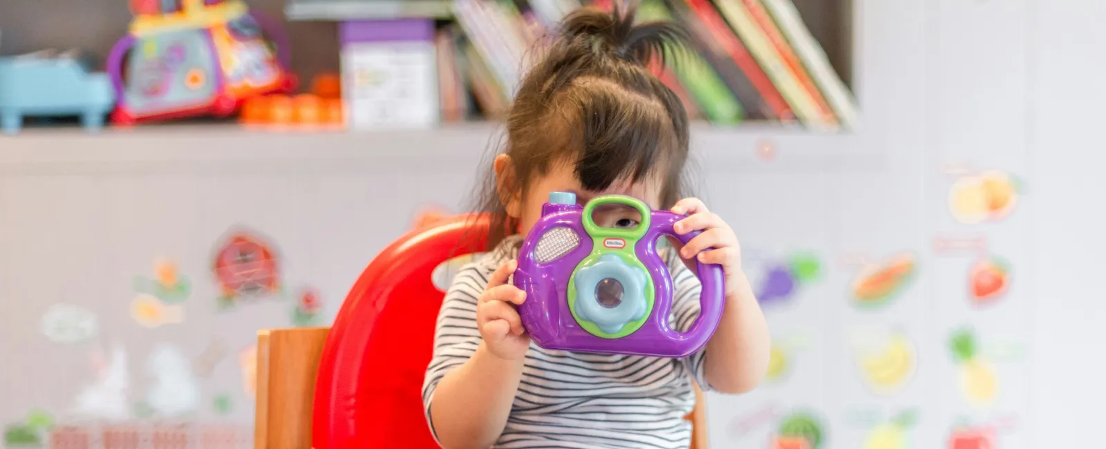 Young child sitting in red chair playing with colorful toy camera in a bright playroom.