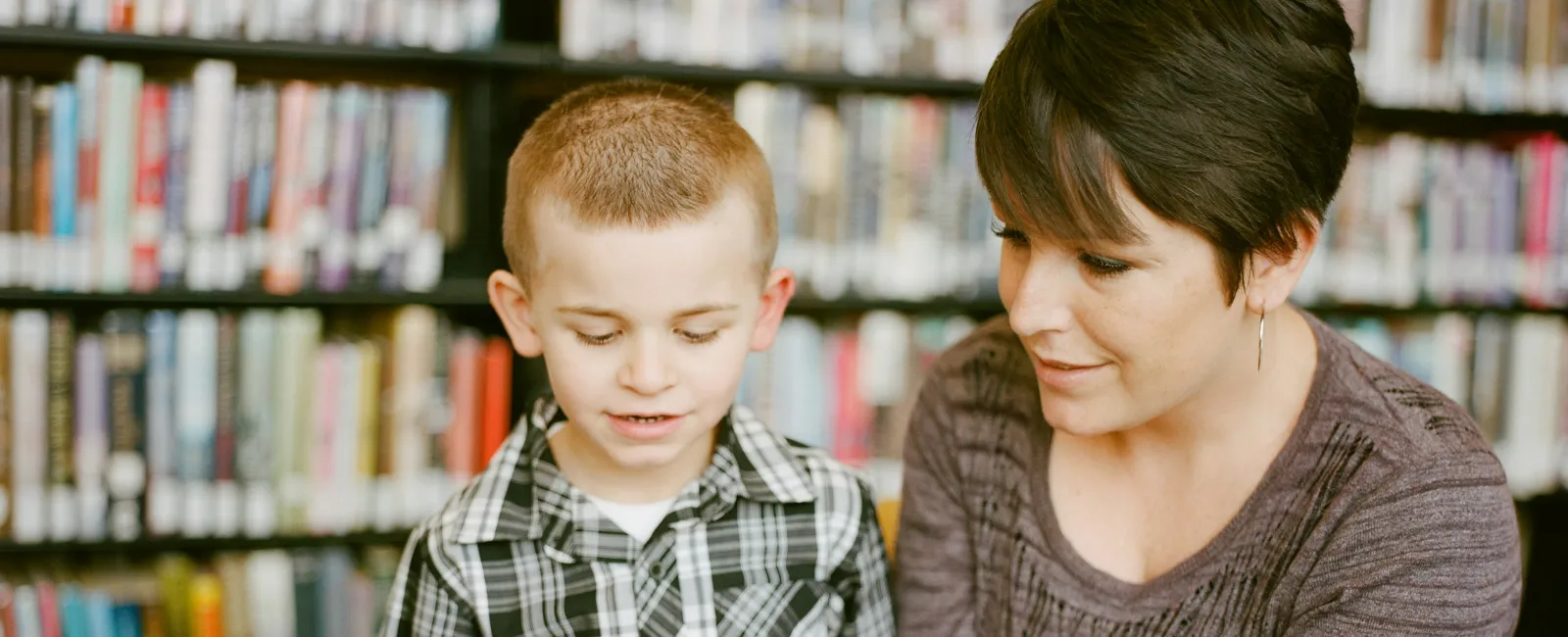 Woman reading a book with a young boy in a library surrounded by bookshelves filled with books