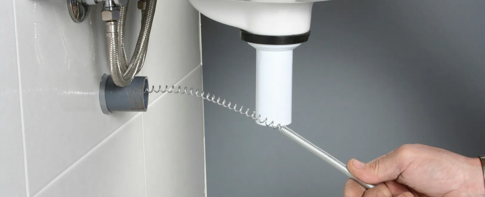 Person using a plumbing snake to clear a clogged sink drain in a modern bathroom setting.