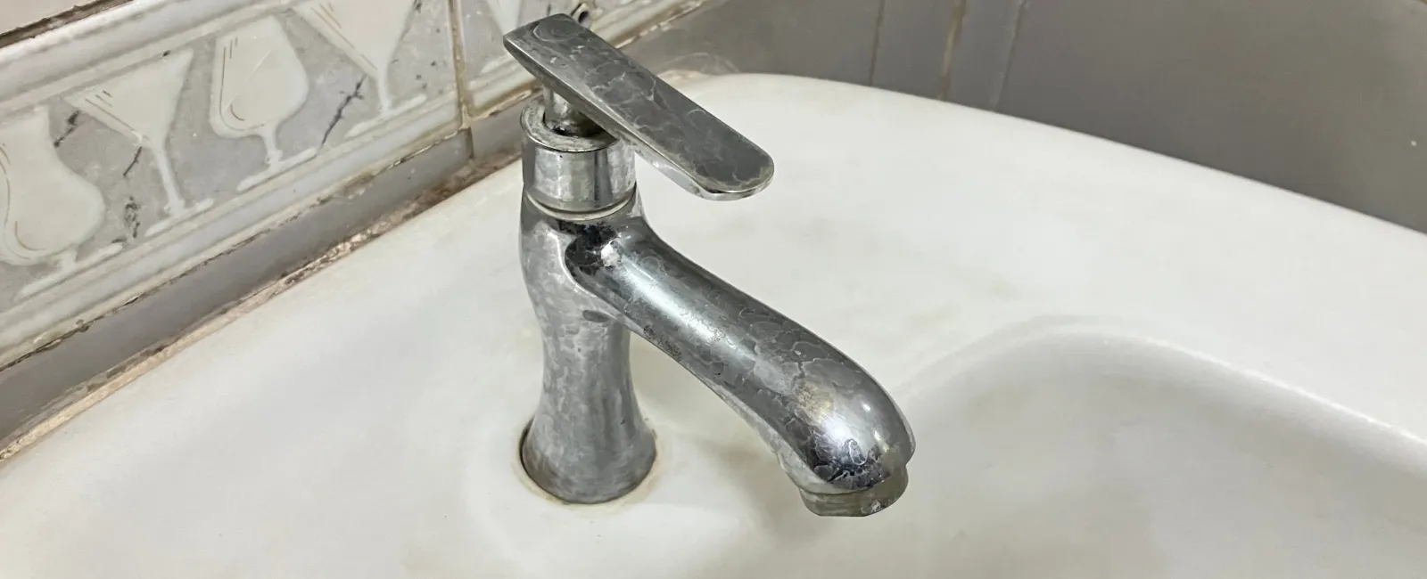 Chrome faucet mounted on a white porcelain sink with decorative patterned tile backsplash in bathroom corner.